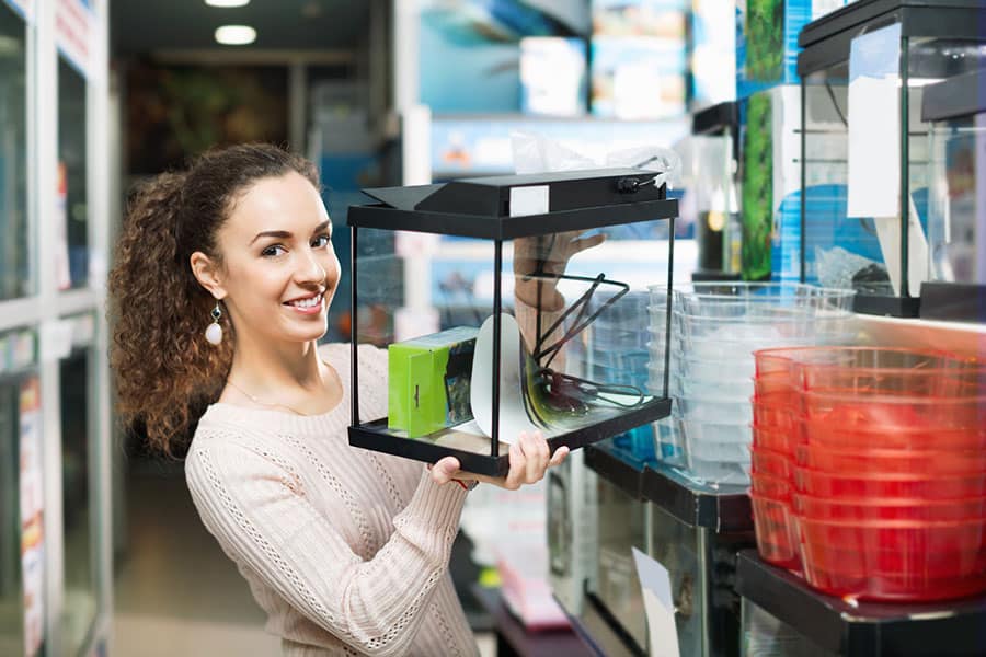 A woman with curly hair, wearing a light sweater, smiles while holding an empty glass aquarium tank with accessories inside at a pet store, surrounded by plastic containers and aquarium supplies.