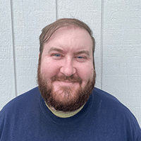 A man with a beard and short hair smiles at the camera, standing in front of a light gray wooden wall. He is wearing a dark blue shirt layered over a light-colored shirt.