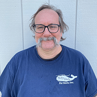 A man with glasses and a bushy mustache is smiling, wearing a navy blue T-shirt with Moby Dick Pet Stores, Inc. printed on it, standing in front of a light gray wooden wall.