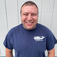 A man with short brown hair smiles broadly while standing in front of a white wooden wall, wearing a navy blue T-shirt with a white logo on the left chest that says Pet Store, Inc.