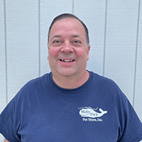 A smiling man with short brown hair wearing a navy blue t-shirt that says Blue Creek Pet Store, Inc. stands in front of a white wooden wall.