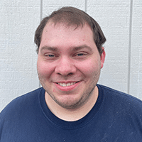 A man with short brown hair and a trimmed beard smiles at the camera, standing in front of a light gray wooden wall. He is wearing a navy blue T-shirt.