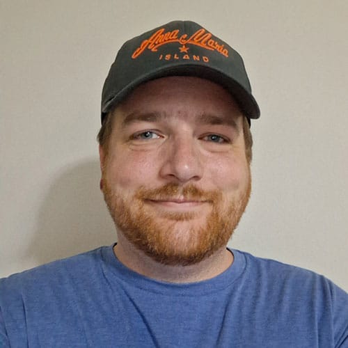 A man with a reddish beard wearing a dark cap with orange text and a blue shirt smiles at the camera against a plain beige background. A man with a reddish beard wearing a dark cap with orange text and a blue shirt smiles at the camera against a plain beige background.