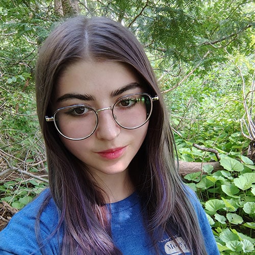 A young person with long brown hair and round glasses is wearing a blue shirt and standing outdoors surrounded by green foliage. A young person with long brown hair and round glasses is wearing a blue shirt and standing outdoors surrounded by green foliage.