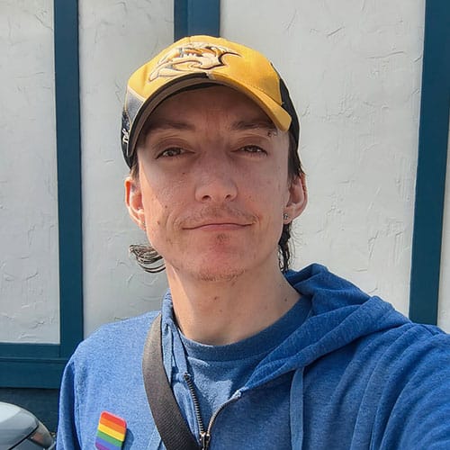 Person wearing a yellow cap with a cartoon logo, blue hoodie, and a rainbow pin, standing outside in sunlight, with a textured white wall and blue trim in the background. Person wearing a yellow cap with a cartoon logo, blue hoodie, and a rainbow pin, standing outside in sunlight, with a textured white wall and blue trim in the background.