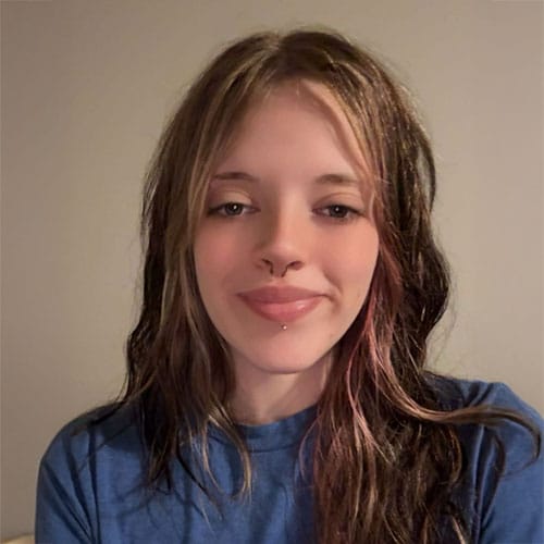 Young person with long, wavy brown hair and blonde highlights, wearing a blue shirt, smiling slightly in front of a plain beige background. Young person with long, wavy brown hair and blonde highlights, wearing a blue shirt, smiling slightly in front of a plain beige background.