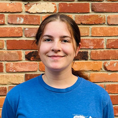 A young person with brown hair tied back, wearing a blue shirt, stands in front of a red brick wall and smiles gently at the camera. A young person with brown hair tied back, wearing a blue shirt, stands in front of a red brick wall and smiles gently at the camera.