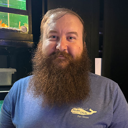 A bearded man in a blue Moby Dick Pet Store shirt stands indoors, smiling slightly. Behind him are fish tanks with green and black frames and some aquarium accessories visible. A bearded man in a blue Moby Dick Pet Store shirt stands indoors, smiling slightly. Behind him are fish tanks with green and black frames and some aquarium accessories visible.
