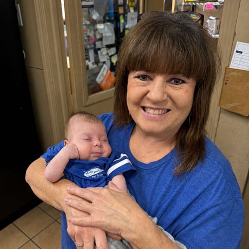 A smiling woman in a blue shirt holds a sleeping baby, also in blue, inside a room with shelves and various items in the background. A smiling woman in a blue shirt holds a sleeping baby, also in blue, inside a room with shelves and various items in the background.
