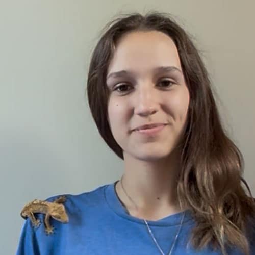 A young woman with long brown hair wearing a blue shirt smiles at the camera. A small brown gecko sits on her left shoulder against a plain light background. A young woman with long brown hair wearing a blue shirt smiles at the camera. A small brown gecko sits on her left shoulder against a plain light background.
