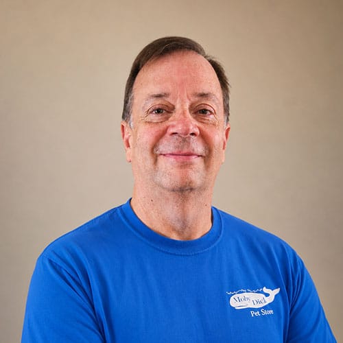 Smiling middle-aged man with short brown hair, wearing a blue Moby Dick Pet Store T-shirt, stands against a plain beige background. Smiling middle-aged man with short brown hair, wearing a blue Moby Dick Pet Store T-shirt, stands against a plain beige background.