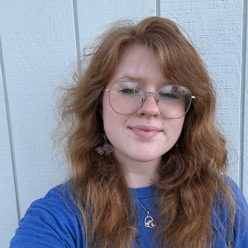 A person with long, wavy reddish-brown hair, wearing glasses and a blue shirt, stands in front of a light-colored wooden wall, smiling slightly at the camera. A person with long, wavy reddish-brown hair, wearing glasses and a blue shirt, stands in front of a light-colored wooden wall, smiling slightly at the camera.